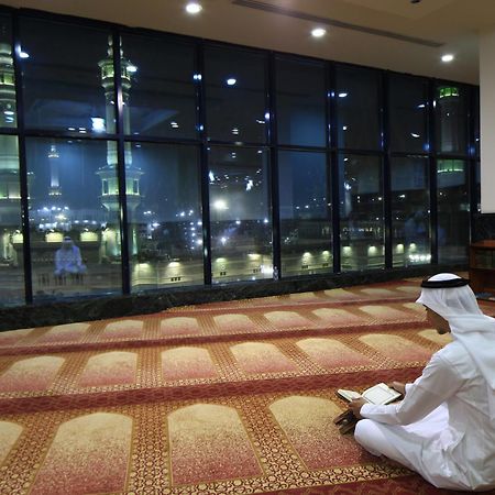 Al Ghufran Safwah Hotel Makkah Mekka Eksteriør billede The photo depicts a person sitting on a carpeted floor, facing away from the camera. They are dressed in traditional attire, likely a white thobe and a headscarf. In the background, large windows reveal a nighttime view of a mosque, with illuminated 