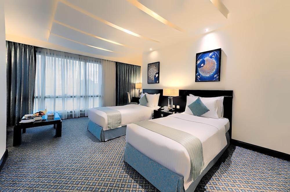 Al Ghufran Safwah Hotel Makkah Mekka Eksteriør billede The photo shows a hotel room with two twin beds. The beds are neatly made with white linens and light blue accents. There are decorative pillows on each bed. A black bedside table with lamps is situated between the beds. On the wall, there are two fr