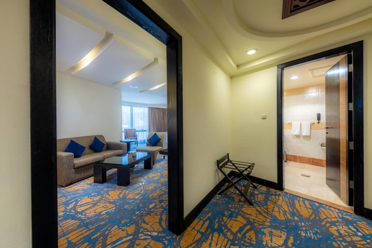 Al Ghufran Safwah Hotel Makkah Mekka Eksteriør billede The photo shows a view of a hotel room entrance. On the left side, there is a seating area with a sofa and a coffee table, decorated with blue cushions. Through a window, natural light illuminates the space. To the right, there is an open door leadin