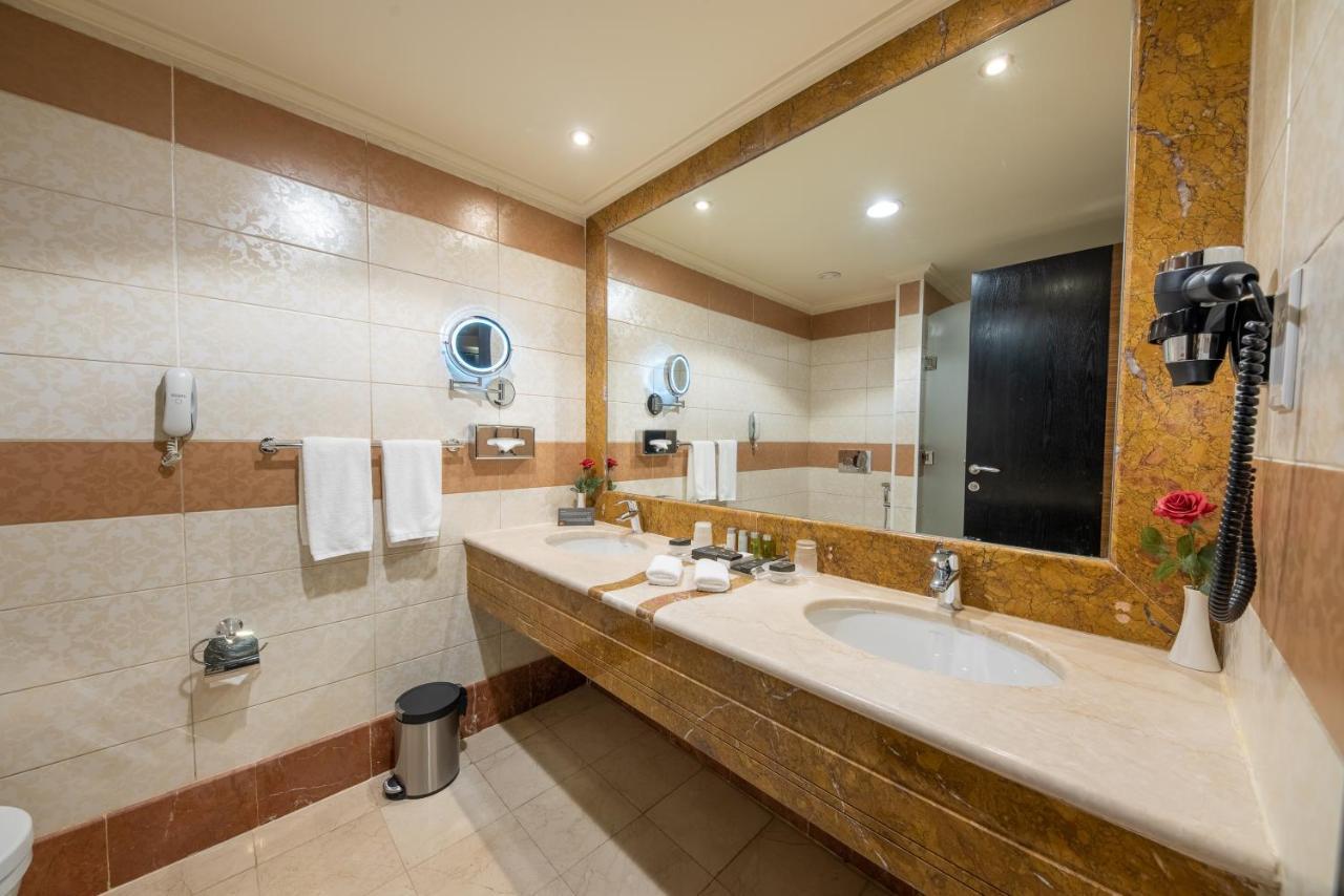 Al Ghufran Safwah Hotel Makkah Mekka Eksteriør billede The photo shows a modern bathroom with a clean and organized design. The walls are tiled with a combination of light and darker colors, and there is a large mirror above a stone countertop. On the countertop, there are toiletries and a small vase wit