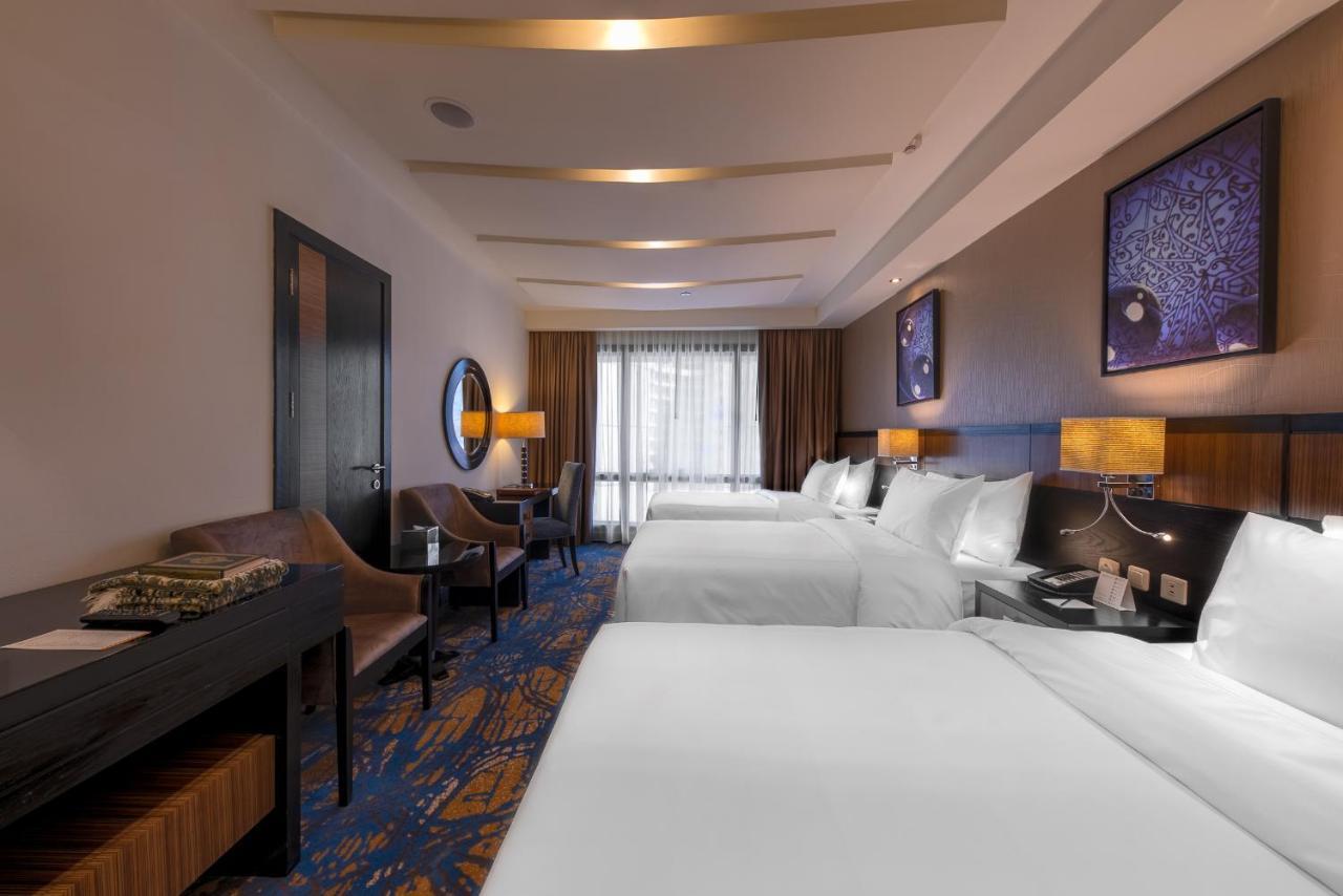 Al Ghufran Safwah Hotel Makkah Mekka Eksteriør billede The photo shows a hotel room featuring two large beds with white bedding. There are two bedside tables with lamps beside each bed. On one side of the room, there is a comfortable chair and a small table, while a work desk can also be seen. The floori