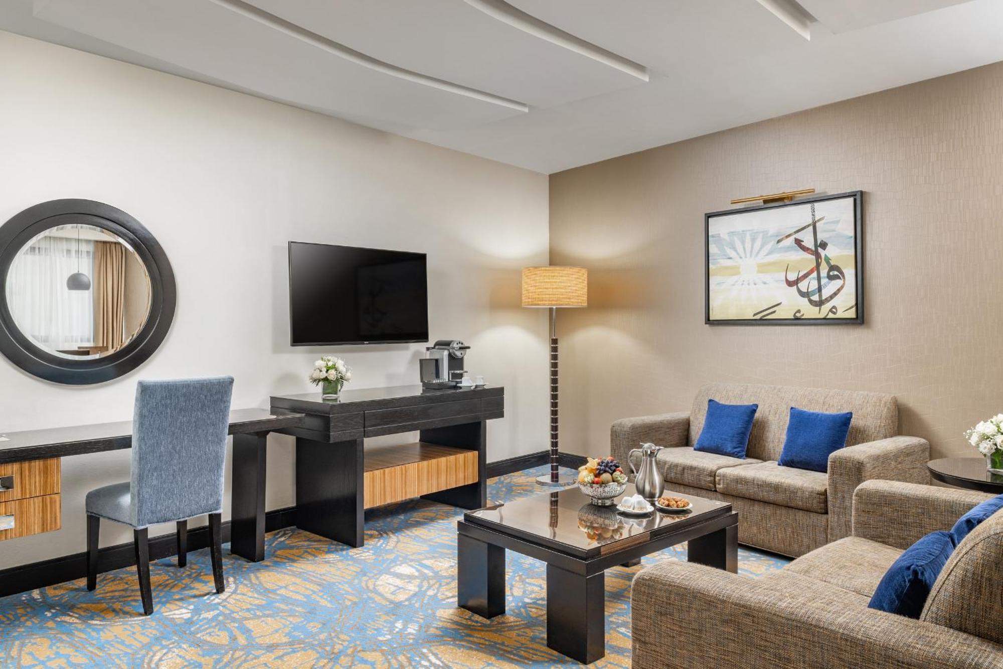 Al Ghufran Safwah Hotel Makkah Mekka Eksteriør billede The photo depicts a stylish hotel room or lounge area. It features a comfortable seating arrangement with two sofas and a coffee table in the center. There is a flat-screen television mounted on the wall, alongside a decorative piece of art. A small 