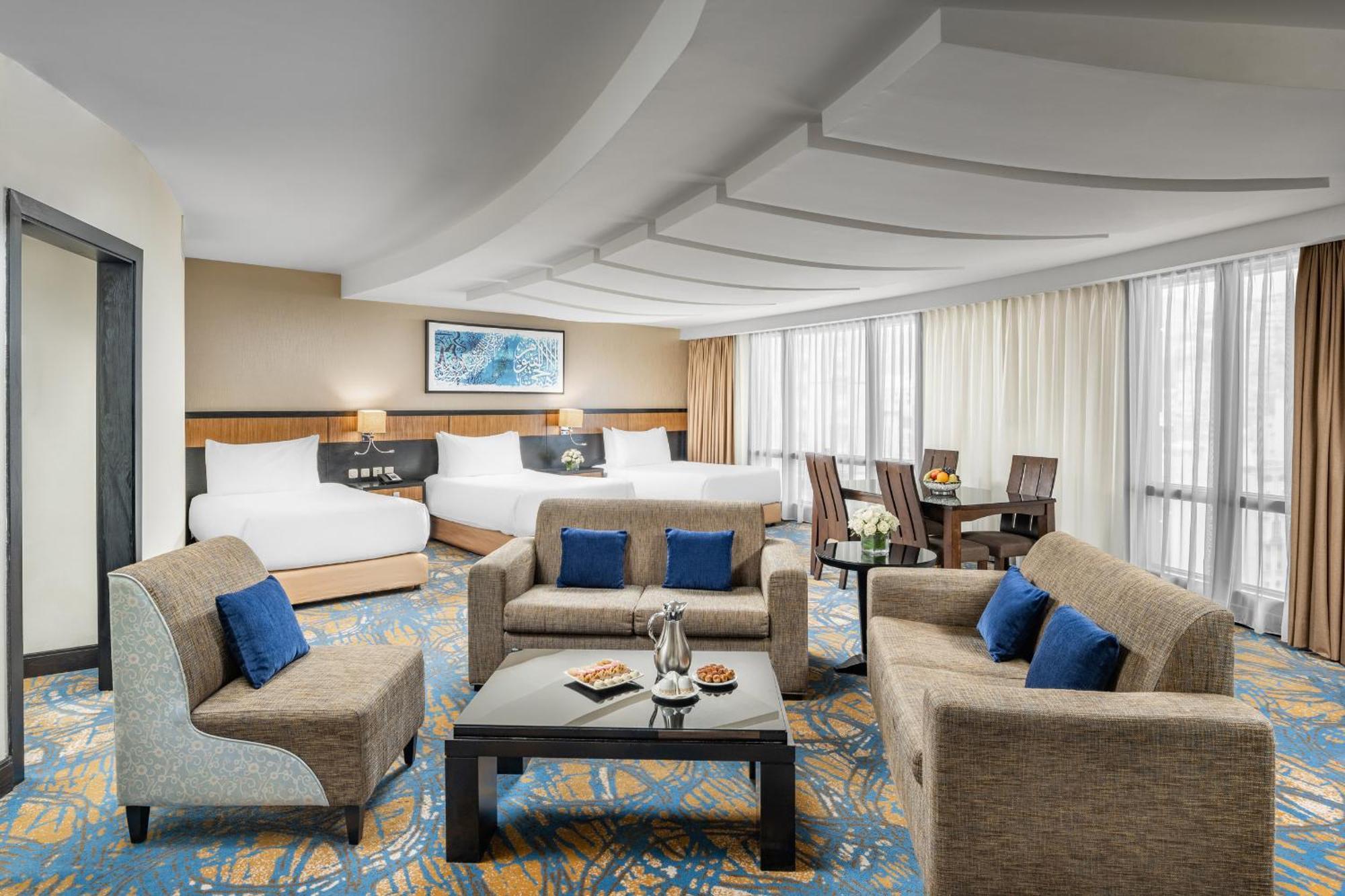 Al Ghufran Safwah Hotel Makkah Mekka Eksteriør billede The photo shows a spacious and elegantly designed hotel room. It features a cozy seating area with a coffee table surrounded by armchairs and a couch, adorned with blue cushions. In the background, there are two beds with white linens, suggesting a c