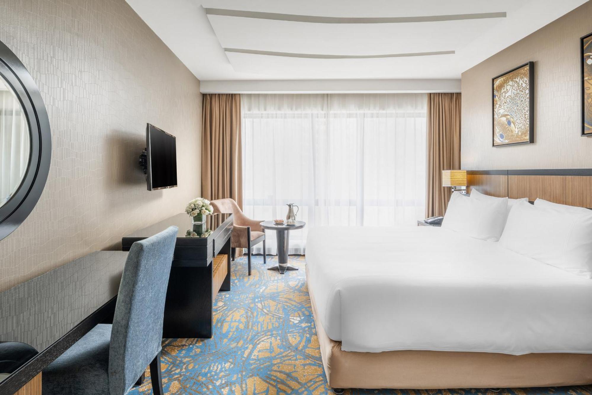 Al Ghufran Safwah Hotel Makkah Mekka Eksteriør billede The photo shows an elegantly designed hotel room. It features a large bed with white linens, a comfy chair, and a small table with two chairs. There is a television mounted on the wall and a decorative piece hanging nearby. The room has large windows