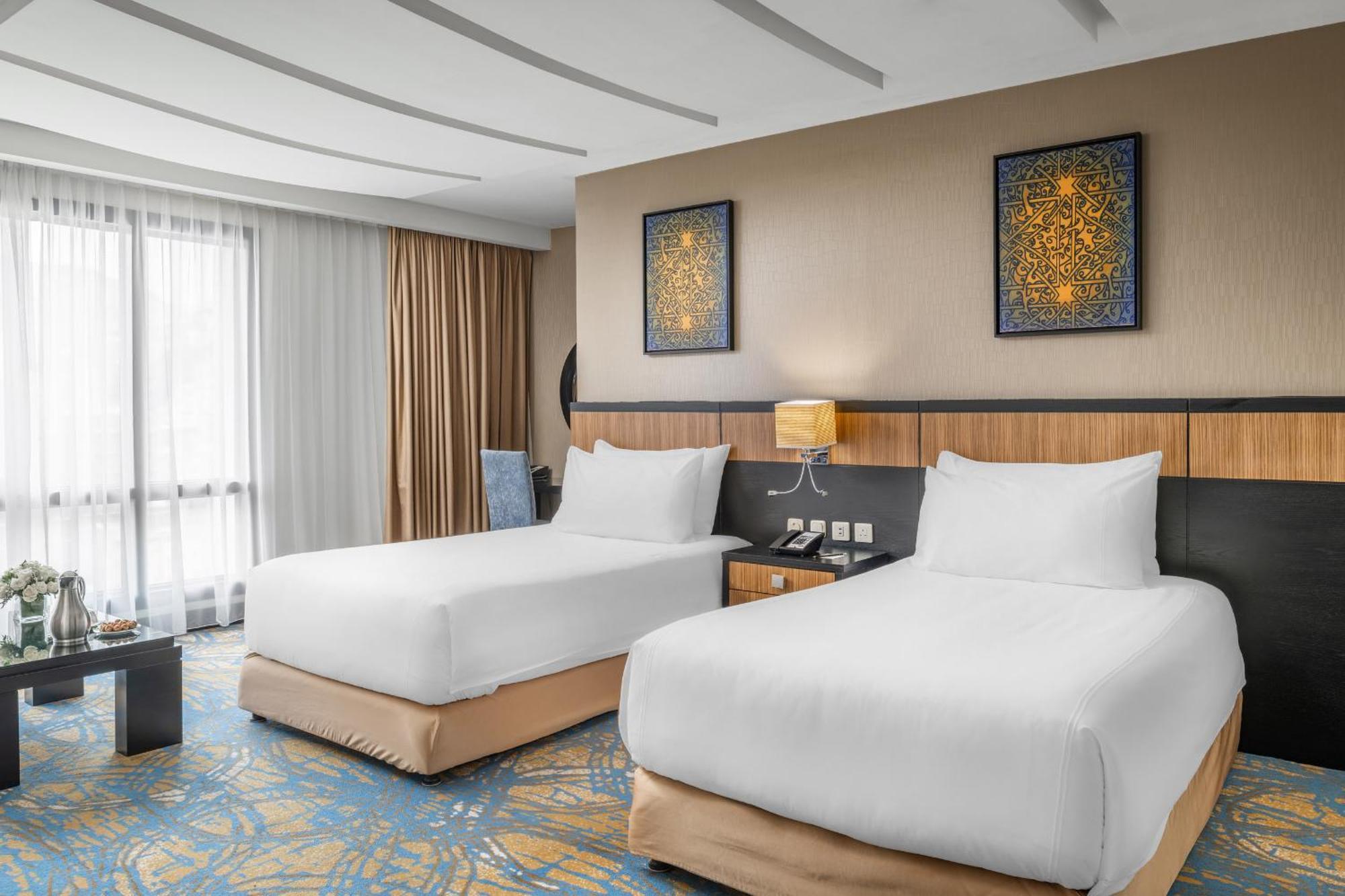 Al Ghufran Safwah Hotel Makkah Mekka Eksteriør billede The photo shows a modern hotel room featuring two white bedding twin beds with soft beige bases. There are decorative paintings on the wall above the beds, and a bedside table with a lamp is positioned between them. The room has large windows covered