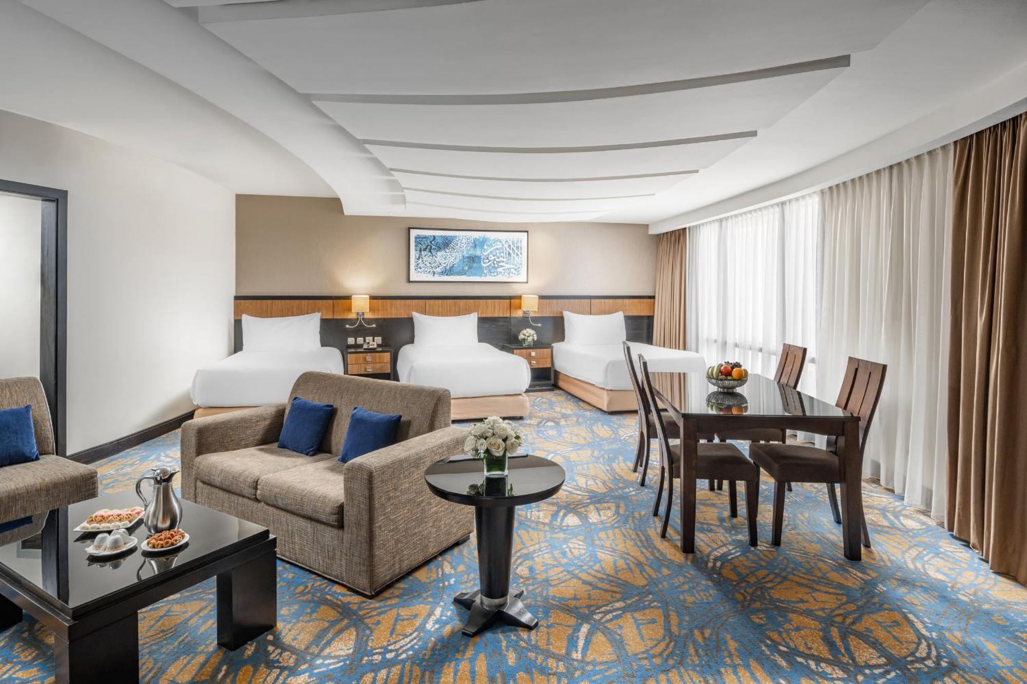 Al Ghufran Safwah Hotel Makkah Mekka Eksteriør billede The photo shows a spacious hotel room designed for comfort. It features two large beds with white bedding, a sitting area with a gray sofa and a coffee table, and a dining area with a wooden table and chairs. There is a decorative vase on the table a